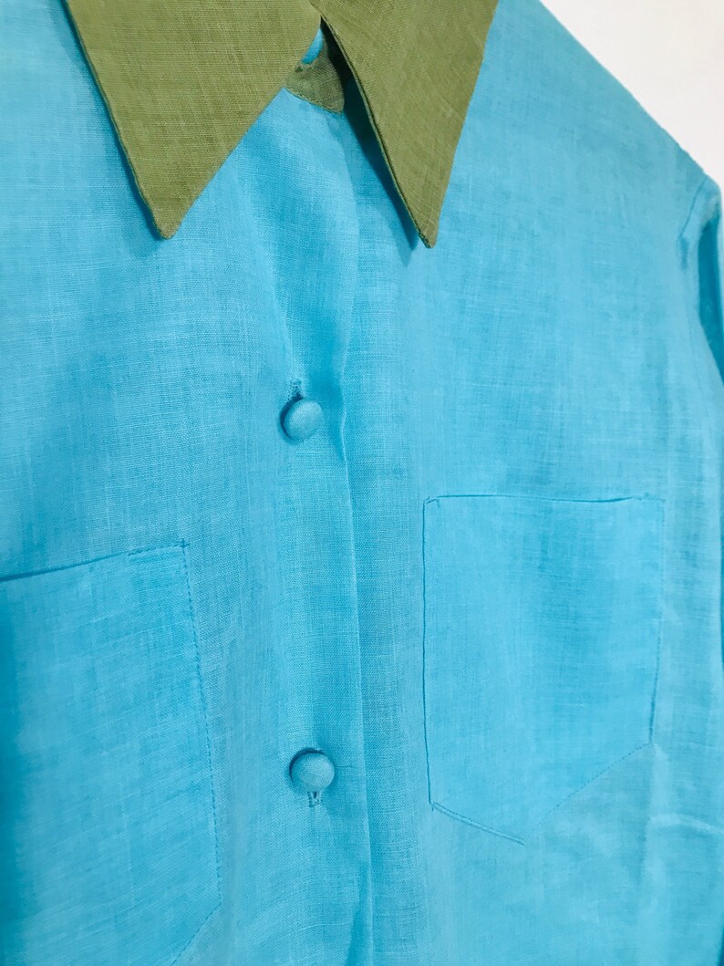 May include: A light blue linen shirt with a green collar and two buttons. The shirt has a pocket on the left side.