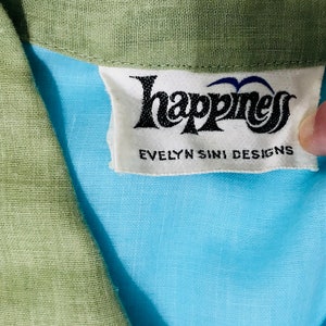 May include: A white tag with black text that reads "Happiness Evelyn Sini Designs". The tag is attached to a light blue fabric with a green fabric collar.