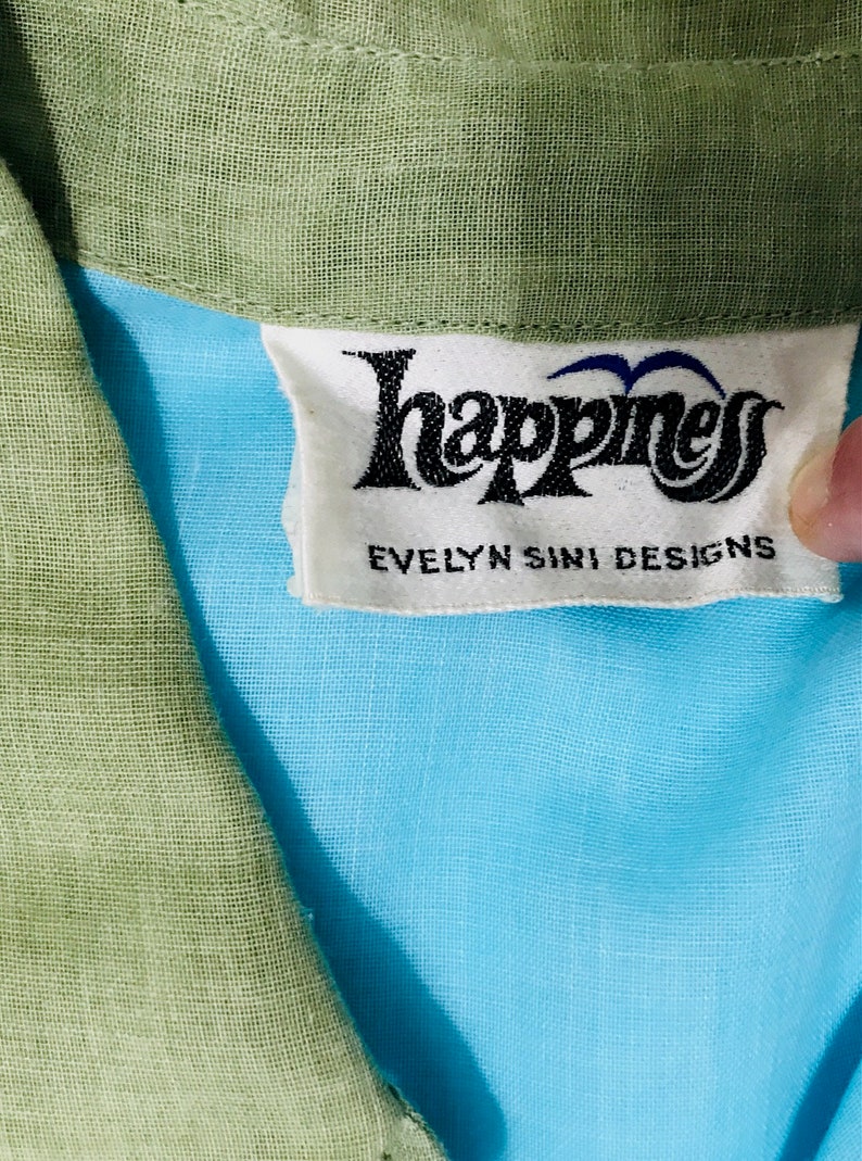 May include: A white tag with black text that reads "Happiness Evelyn Sini Designs". The tag is attached to a light blue fabric with a green fabric collar.
