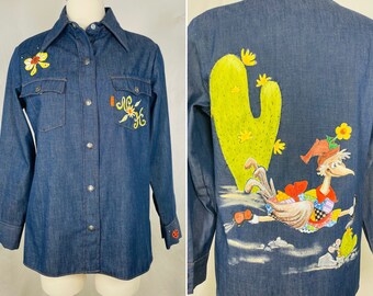 May include: A blue denim shirt with a hand-painted design on the back. The design features a cartoon bird with a red hat, a green cactus, and a yellow flower. The front of the shirt has embroidered flowers and a small embroidered design.