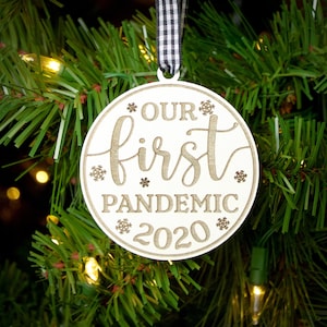 May include: White wooden Christmas ornament with the text "Our first pandemic 2020" in gold lettering. The ornament is decorated with snowflakes.