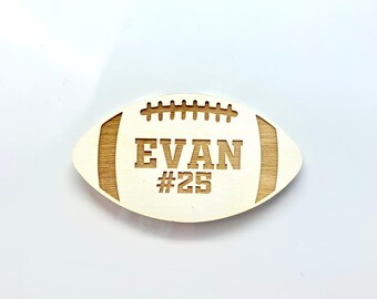 May include: Wooden football-shaped ornament with the name "Evan" and the number "#25" engraved on it.