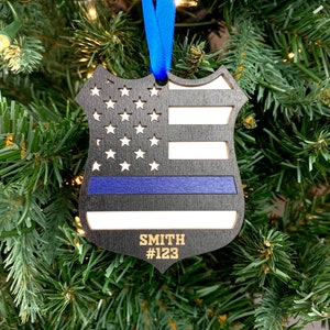 May include: Black wooden ornament shaped like a police badge with a thin blue line and an American flag design. The ornament is personalized with the name "SMITH" and the number "#123".