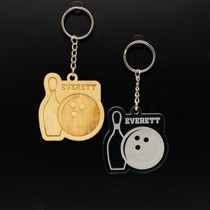 May include: Two keychains with a bowling pin and ball design. One keychain is made of wood and the other is made of clear acrylic. Both keychains have the name "EVERETT" engraved on them.