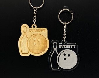 May include: Two keychains featuring a bowling pin and ball design. One keychain is made of wood and the other is made of clear acrylic. Both keychains have the name "EVERETT" engraved on them.