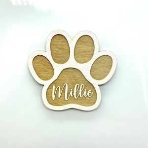 May include: A wooden paw print with a white outline and the name "Millie" written inside. The paw print is a light brown color.