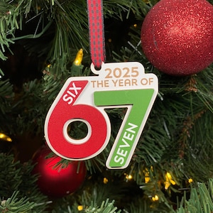 May include: A Christmas tree ornament with the text "2025 The Year of" and the numbers "67" in red and green. The ornament is hanging on a Christmas tree with red and green ornaments.