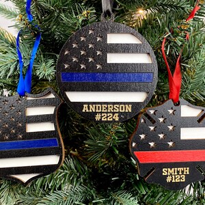May include: Three wooden Christmas ornaments with a thin blue line, thin red line, and American flag designs. The ornaments are personalized with names and numbers.  "Anderson #224", "Smith #123"