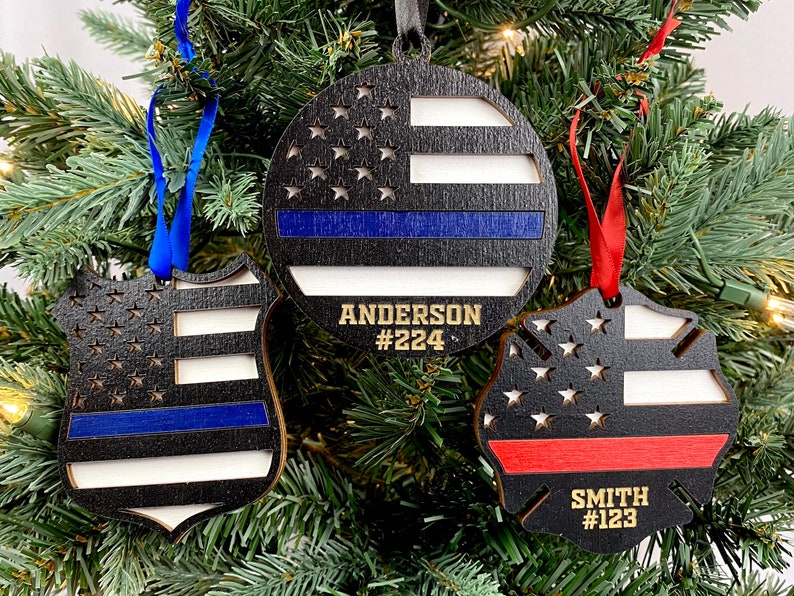 May include: Three wooden Christmas ornaments with a thin blue line, thin red line, and American flag designs. The ornaments are personalized with names and numbers.  "Anderson #224", "Smith #123"