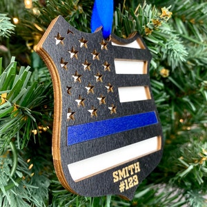 May include: A wooden ornament shaped like a police badge with an American flag design and a blue line. The ornament is personalized with the name "SMITH" and the number "#123".