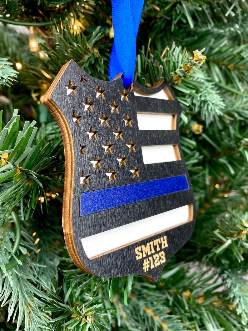 May include: A wooden ornament shaped like a police badge with an American flag design and a blue line. The ornament is personalized with the name "SMITH" and the number "#123".
