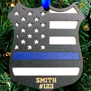 May include: Black wooden ornament shaped like a police badge with an American flag design and a blue stripe. The ornament has the name "SMITH" and the number "#123" engraved on it.