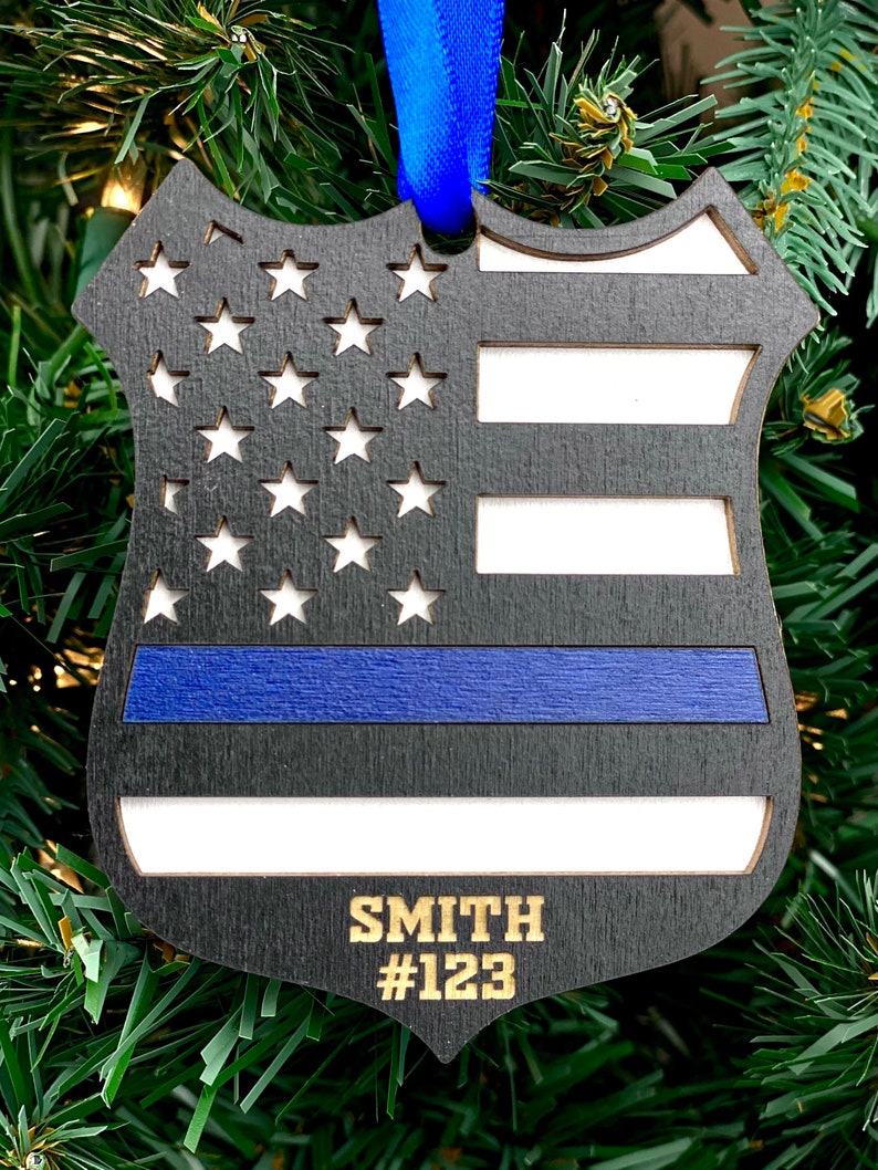 May include: Black wooden ornament shaped like a police badge with an American flag design and a blue stripe. The ornament has the name "SMITH" and the number "#123" engraved on it.