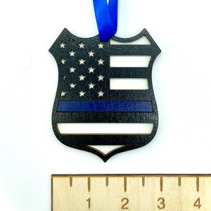 May include: Wooden ornament shaped like a police badge with an American flag design and a blue stripe across the middle. The ornament is approximately 3 inches tall.