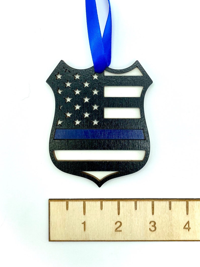 May include: Wooden ornament shaped like a police badge with an American flag design and a blue stripe across the middle. The ornament is approximately 3 inches tall.