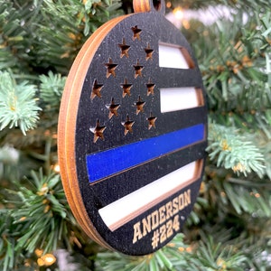 May include: A round wooden ornament with a black and white American flag design and a blue thin blue line. The ornament has the name "ANDERSON #224" written on it in gold lettering.