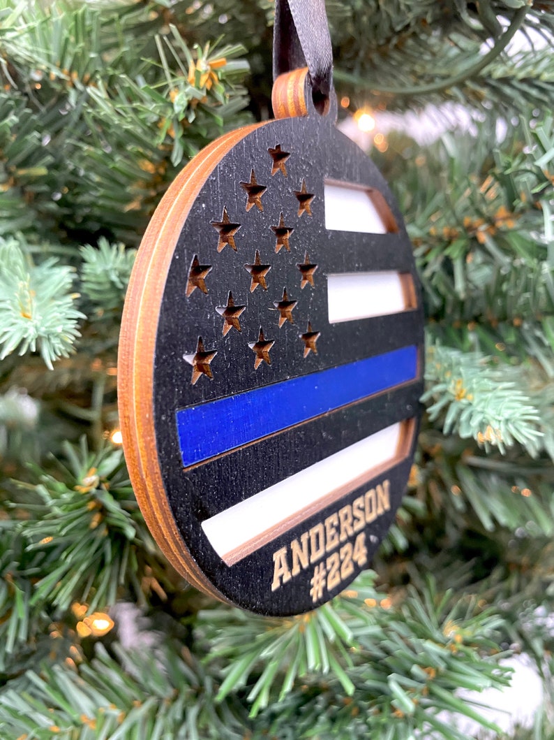 May include: A round wooden ornament with a black and white American flag design and a blue thin blue line. The ornament has the name "ANDERSON #224" written on it in gold lettering.
