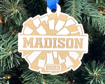 May include: Wooden Christmas ornament with the name "MADISON" engraved in a rectangular banner. The ornament has a cheerleader design with the year "2023" and a blue ribbon for hanging. The ornament is hanging on a Christmas tree.