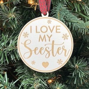 May include: White wooden Christmas ornament with the text "I love my Seester" in gold lettering. The ornament is decorated with snowflakes and a small heart.