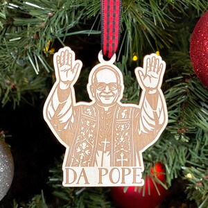 May include: Wooden Christmas ornament featuring a depiction of a man with his arms raised. The ornament has the text "DA POPE" at the bottom. It hangs from a red and black ribbon against a backdrop of a Christmas tree and ornaments.