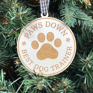 May include: White wooden Christmas ornament with the text "Paws Down Best Dog Trainer" and a brown paw print in the center. The ornament is hanging on a green Christmas tree.