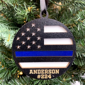 May include: A round wooden ornament with a black and white American flag design and a blue stripe across the middle. The ornament has the name "Anderson #224" written in gold lettering.