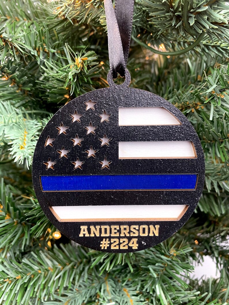May include: A round wooden ornament with a black and white American flag design and a blue stripe across the middle. The ornament has the name "Anderson #224" written in gold lettering.