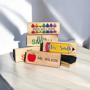 May include: A set of five wooden teacher nameplates with different designs. The top nameplate features a row of colorful crayons and the text "MISS WILLIAMS". The second nameplate has the text "Mrs. SMITH" in green letters. The third nameplate has a pencil design and the text "Mrs. Smith". The fourth nameplate has a red apple design and the text "MR. WILSON". The fifth nameplate has a black chalkboard eraser.