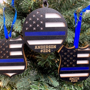 May include: Three wooden Christmas ornaments with a thin blue line flag design. The ornaments are shaped like a circle, a shield, and a badge. The ornaments are personalized with names and badge numbers. The ornaments are hanging on a Christmas tree.