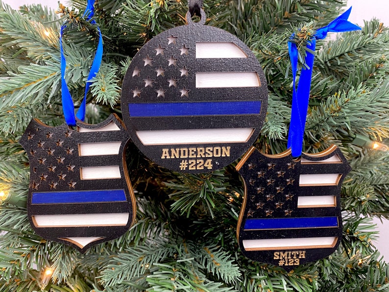 May include: Three wooden Christmas ornaments with a thin blue line flag design. The ornaments are shaped like a circle, a shield, and a badge. The ornaments are personalized with names and badge numbers. The ornaments are hanging on a Christmas tree.