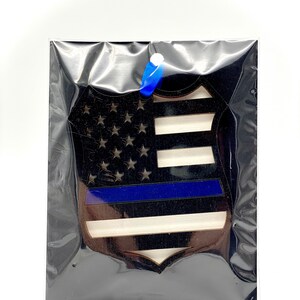 May include: A wooden ornament shaped like a police badge with an American flag design and a blue line across the middle.