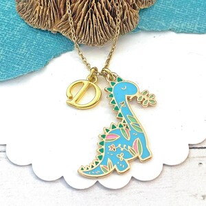 May include: Gold necklace with a blue dinosaur pendant, decorated with pink and green floral details, and a gold letter "D" charm. The dinosaur pendant is on a gold chain. The necklace is displayed on a white, scalloped edge surface.