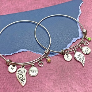 May include: Two silver bangle bracelets with charms. The charms include a heart, a letter W, the word "Best", the word "BFF", a letter G, the word "Friends", and a heart with a swirl design. The bracelets are on a pink surface.