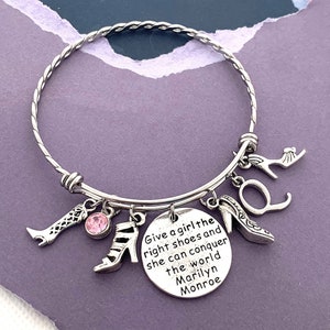 May include: Silver charm bracelet with a quote from Marilyn Monroe: "Give a girl the right shoes and she can conquer the world". The bracelet features several shoe charms, including a high heel, a boot, and a small pink rhinestone.
