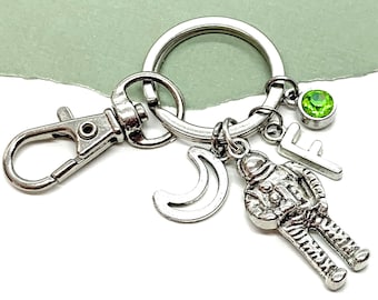 May include: Silver keychain with a silver astronaut charm, a crescent moon charm, a letter "F" charm, and a green gemstone charm.
