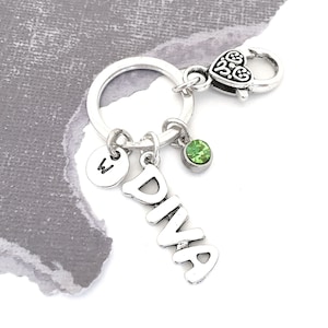 May include: Silver-toned keychain with a round key ring and a heart-shaped clasp. The keychain features the word "DIVA" in a vertical font, a small green gemstone, and a circular charm with the letter "W".