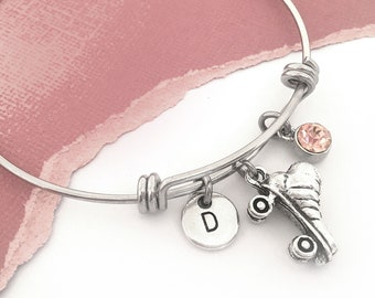 May include: A silver-colored bangle bracelet with a charm featuring a roller skate, a round charm with the letter "D", and a small pink gemstone. The bracelet is displayed on a pink background.