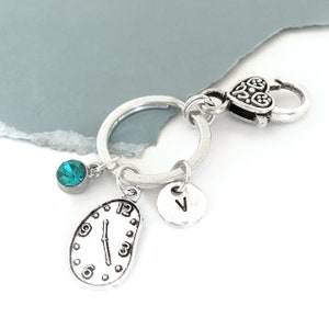 May include: Silver keychain with a melted clock charm, a teal blue crystal charm, and a silver charm with the letter "V" on it. The keychain has a silver heart-shaped clasp.