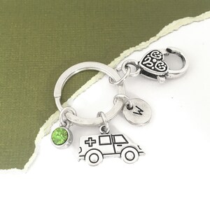 May include: Silver keychain with a green crystal charm, a silver ambulance charm, and a silver charm with the letter "M" on it. The keychain has a silver heart-shaped clasp.