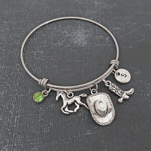 May include: A silver expandable bangle bracelet with charms including a horse, a cowboy hat, a boot, a green gemstone, and a silver charm with the letter "S".