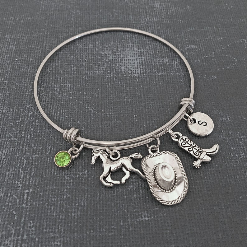 May include: A silver expandable bangle bracelet with charms including a horse, a cowboy hat, a boot, a green gemstone, and a silver charm with the letter "S".