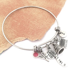 Op de afbeelding: Een zilveren armband met verschillende bedels, waaronder een vingerhoed, een schaar, een spoel garen en een bedel met de tekst "Silk Cord".