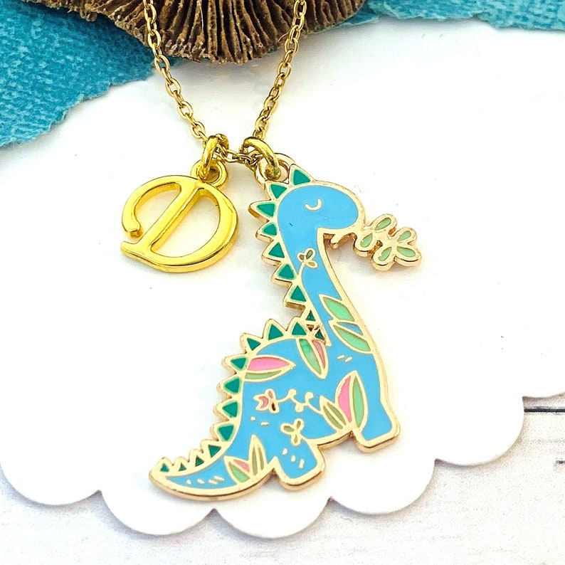 May include: A gold-coloured necklace featuring a pendant with a light blue dinosaur design, accented with green and pink floral details. The necklace also includes a gold letter "D" charm. The dinosaur is holding a sprig of leaves.