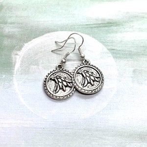 May include: Silver dangle earrings with a circular design featuring an eagle head in relief.