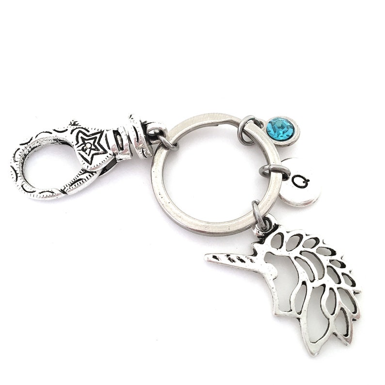Puede incluir: Llavero plateado con un dije de unicornio, un dije de cristal azul y un dije plateado con la letra 'G'.