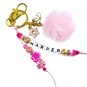 May include: A pink and gold keychain with a fluffy pink pom-pom, daisy and star charms, and beads spelling "HARPER". The keychain has a gold clasp and various pink beads and charms.