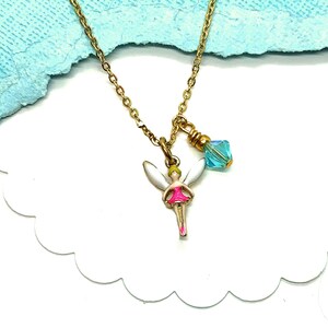 May include: A gold-colored necklace with a fairy pendant and a blue crystal charm. The fairy pendant has white wings, a pink dress, and a yellow head. The necklace is on a white surface with a light blue background.