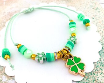May include: A green four-leaf clover charm hangs from a green and white beaded bracelet. The bracelet is made with a green cord and has a variety of green and white beads, including hearts and flowers.
