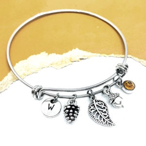 May include: A silver expandable bangle bracelet with charms. The charms include a small acorn, a leaf, a pine cone, and a round charm with the letter "W" on it.