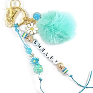 May include: A personalized keychain with the name "Shelby" spelled out in white letter beads. The keychain features a gold clasp, a fluffy turquoise pom-pom, and decorative beads in shades of blue, white, and gold. Charms include a daisy and a smiling face.